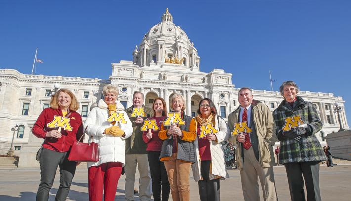 Members of the Board of  holding a block M sign and standing in front of the state capitol